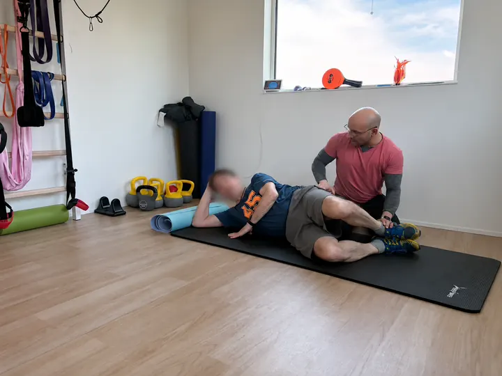 Samuel supports a client's hip and ankle during a side-lying hip mobility drill on an exercise mat.